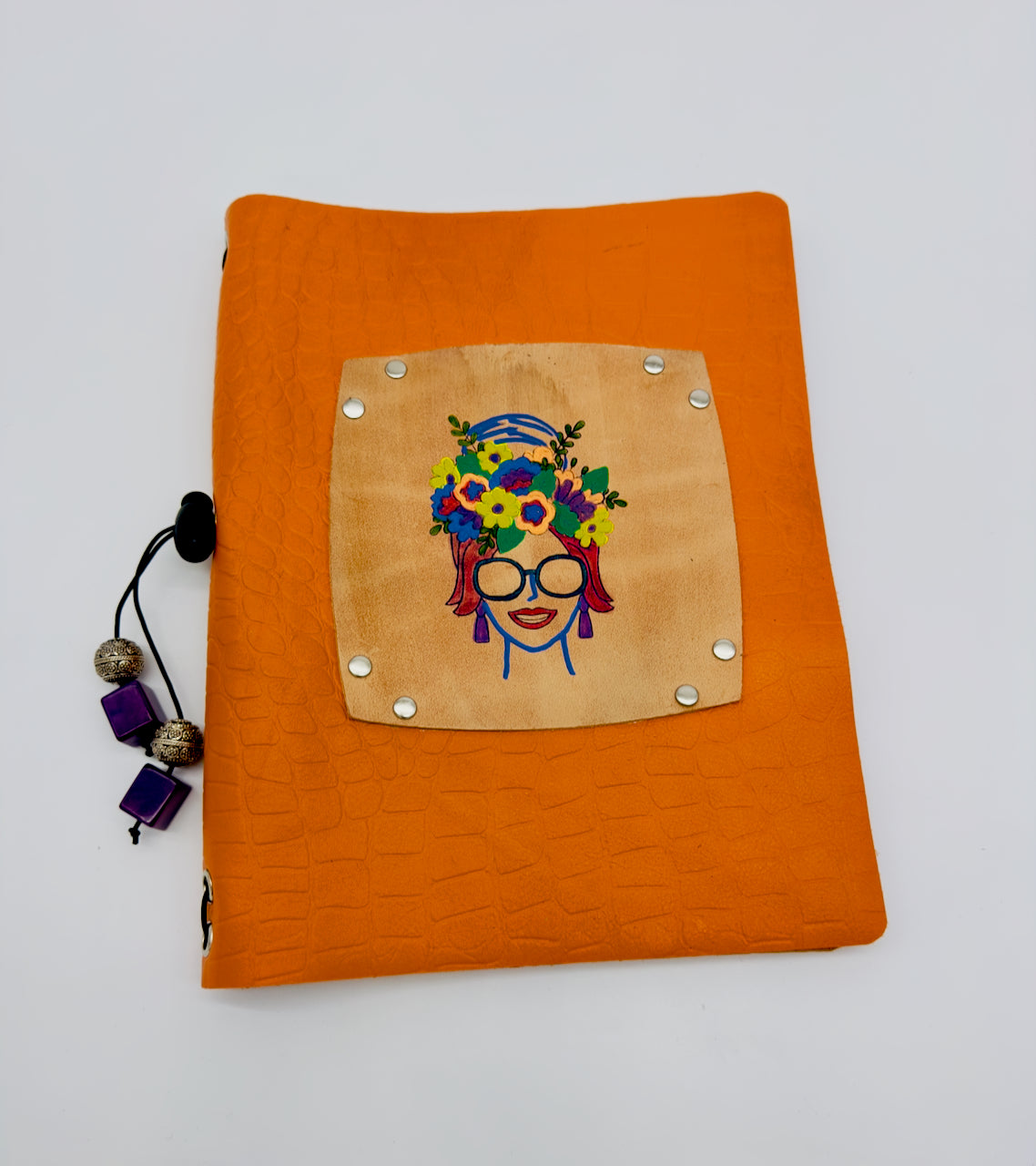 An orange leather journal with a crocodile print pattern and a decorative floral lady design on the cover. It includes silver and purple beads attached to an elastic band for journal replacement.