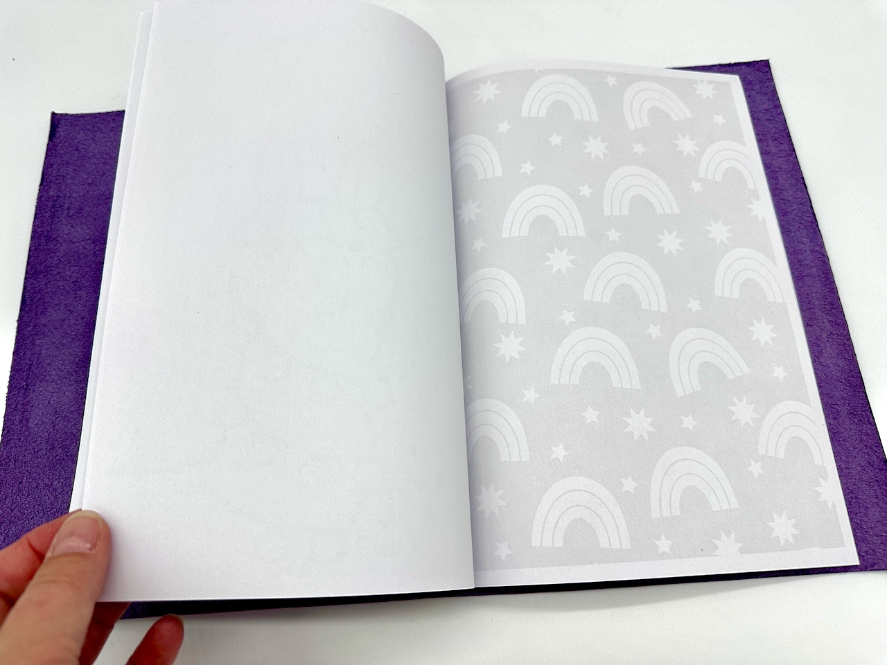 Open leather journal book with purple leather and inside notebook paper page with rainbow images, held by a hand.