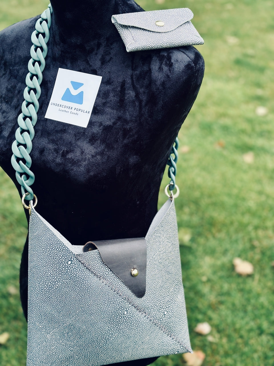 Grayish green shagreen leather crossbody handbag with chain strap and matching wallet on a mannequin against a grassy background