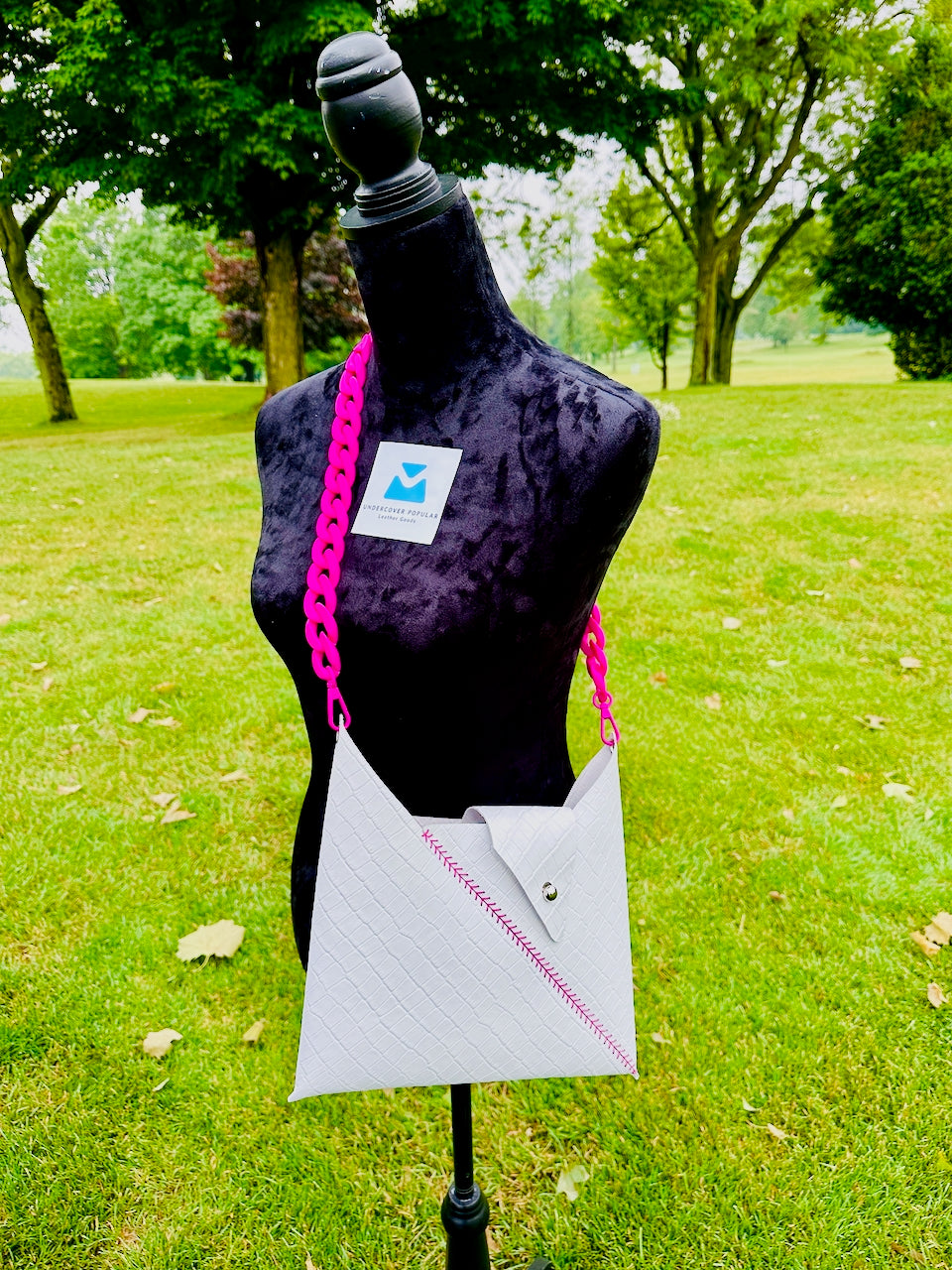 White crocodile embossed leather crossbody handbag with pink chain strap on a mannequin against a green outdoor background