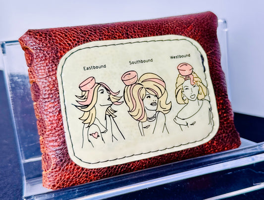 leather wallet with a graphic of three women labeled 'Eastbound', 'Southbound', and 'Westbound' on a clear stand.