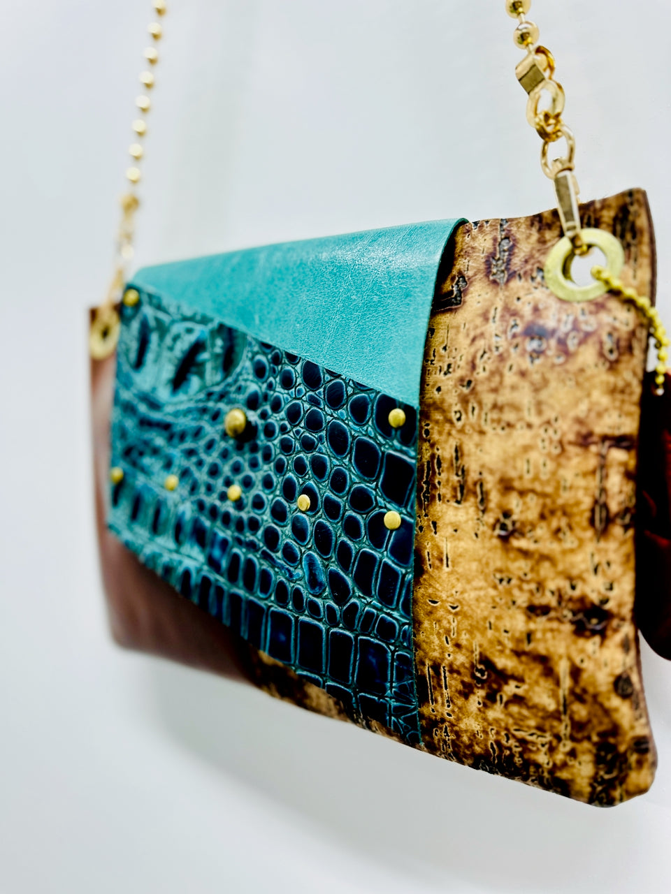 A handmade leather patchwork shoulder bag featuring abrown and textured cork panels, a solid turquoise and black and teal crocodile printed leather flap and gold-tone studs and chain strap.