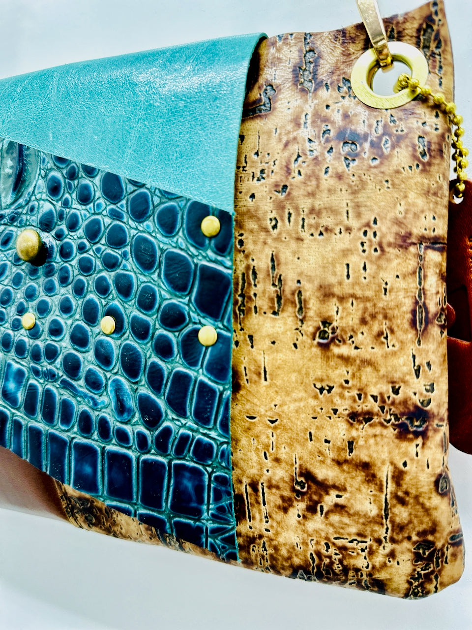 A close-up of a handmade shoulder purse featuring a split design: one side has a glossy turquoise crocodile-embossed texture with small gold studs, and the other side has a distressed tan-brown cork leather panel with a gold ring and chain detail.