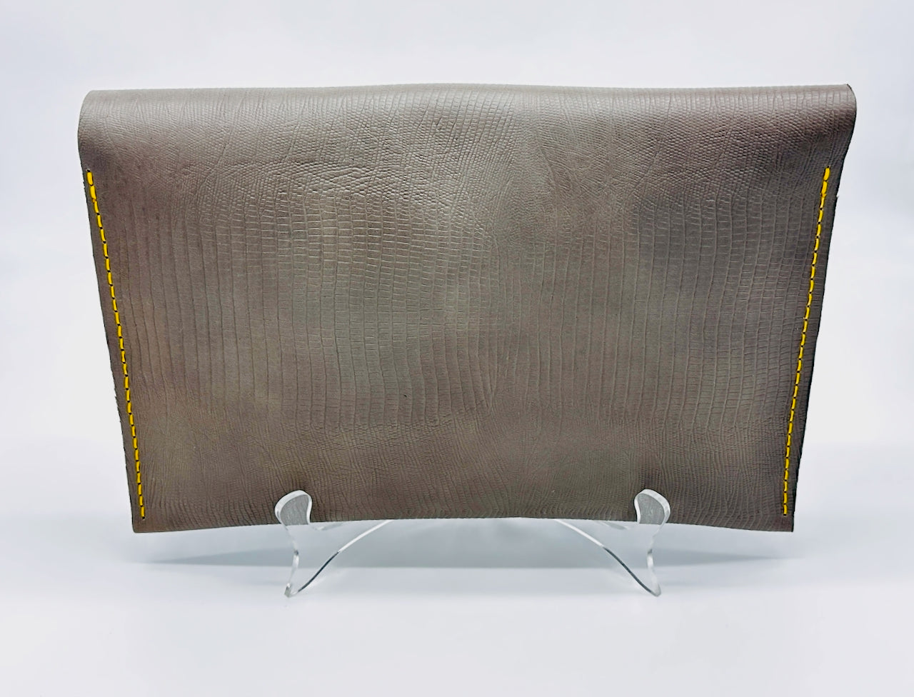 Back side of a Gray leather clutch with yellow stitching on an acrylic easel against a white background