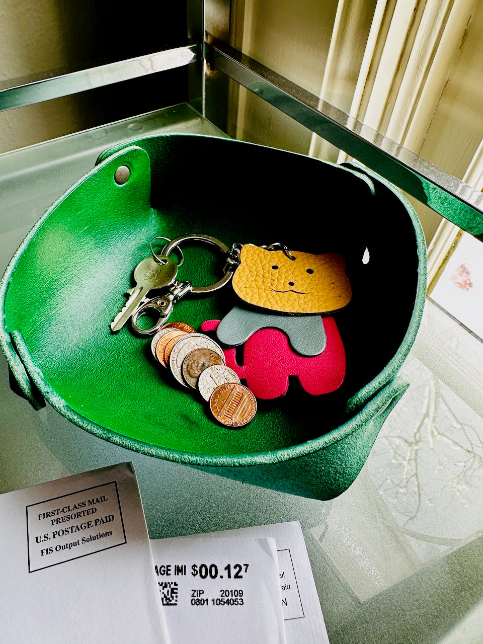Rounded square leather tray holds coins and keychain near mail on glass shelf