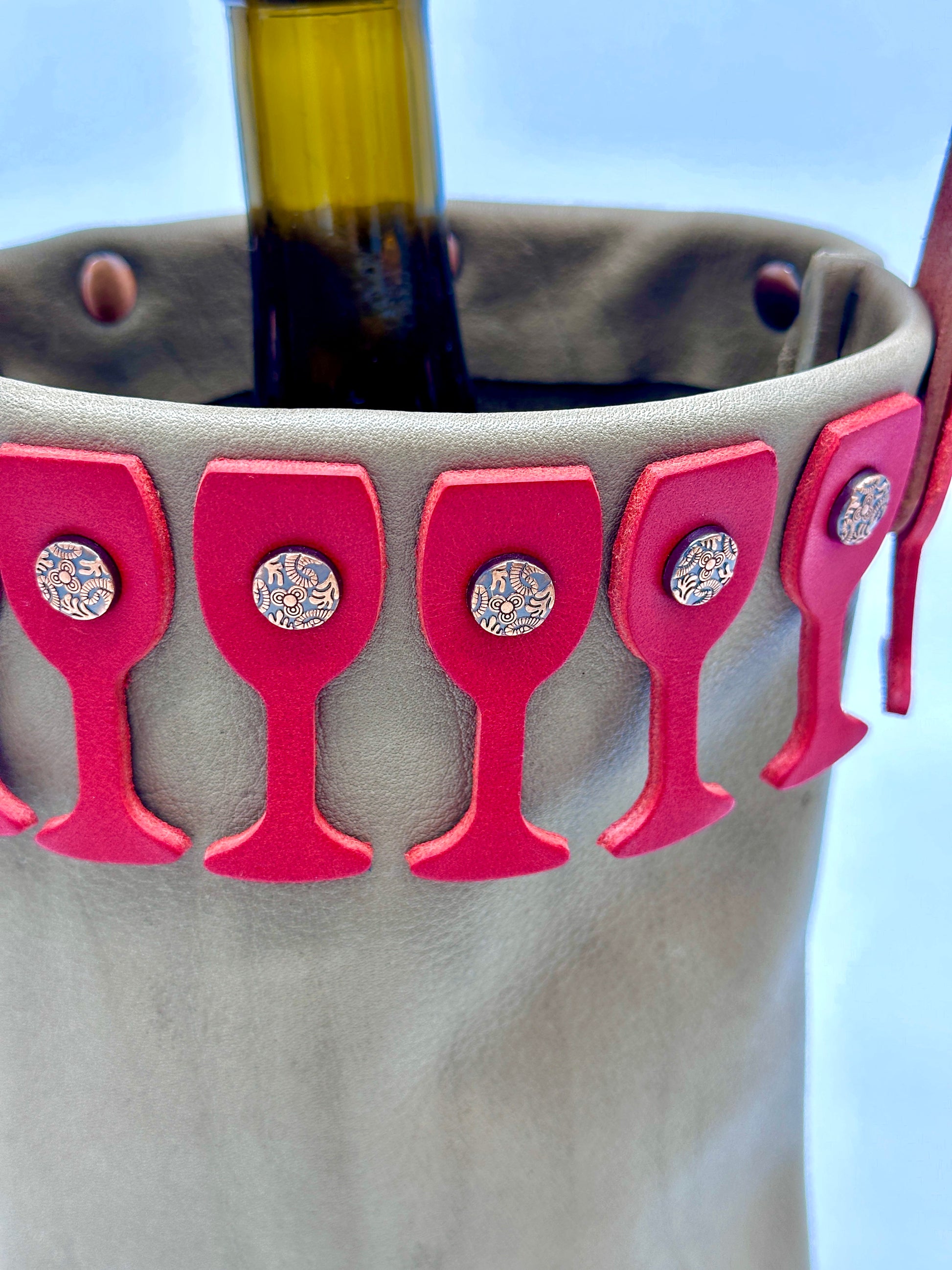 Beige leather wine bottle holder with red wine glass cutouts and decorative copper rivets.