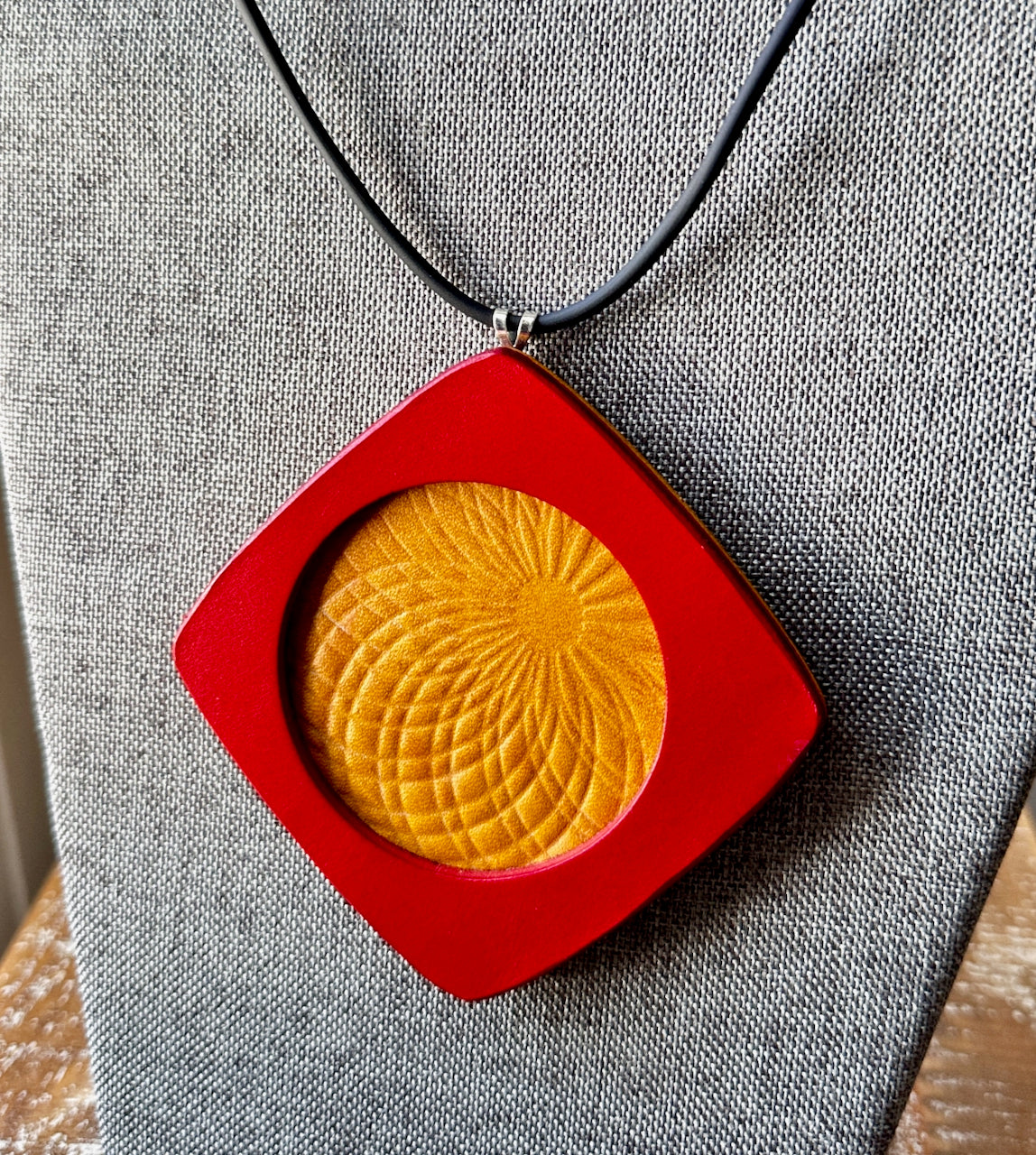 Necklace with a red square leather pendant featuring a yellow circular embossed sun flower design on a gray fabric background