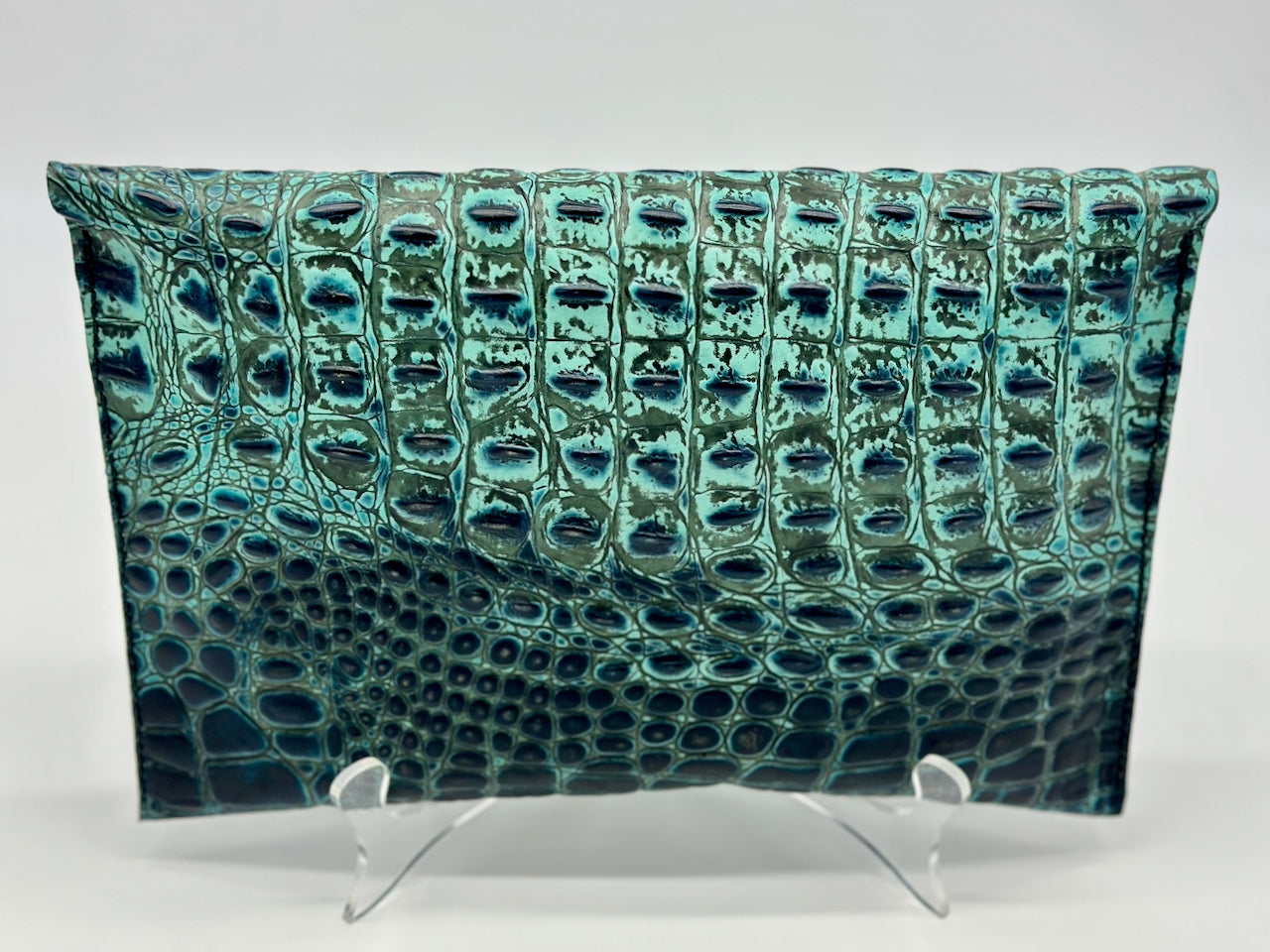 Teal clutch bag with crocodile texture on a stand against a white background