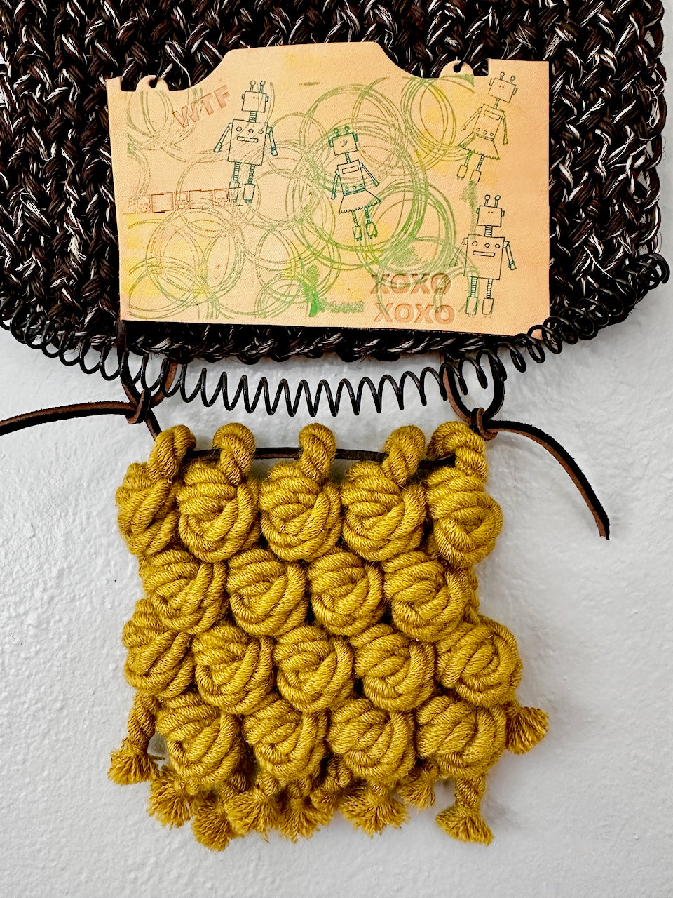 Yellow knitted wall hanging with a stamped leather patch featuring robot illustrations on a textured surface.