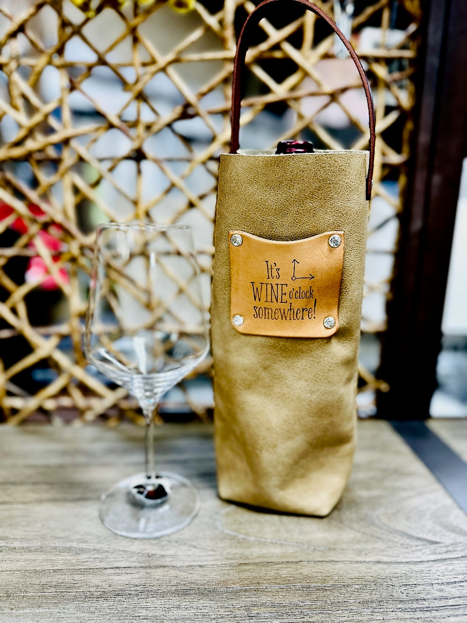Leather wine bottle carrier with leather patch that reads "its wine o'clock somewhere!" and wine glass on table