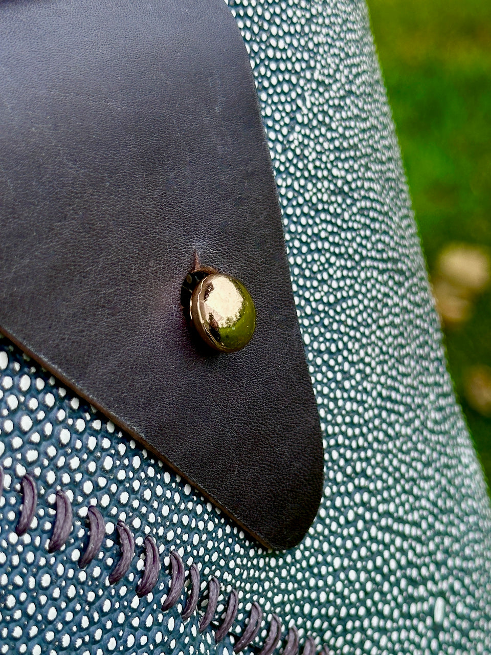 Close-up of a gold button on a textured dark green shagreen leather surface of a crossbody purse.