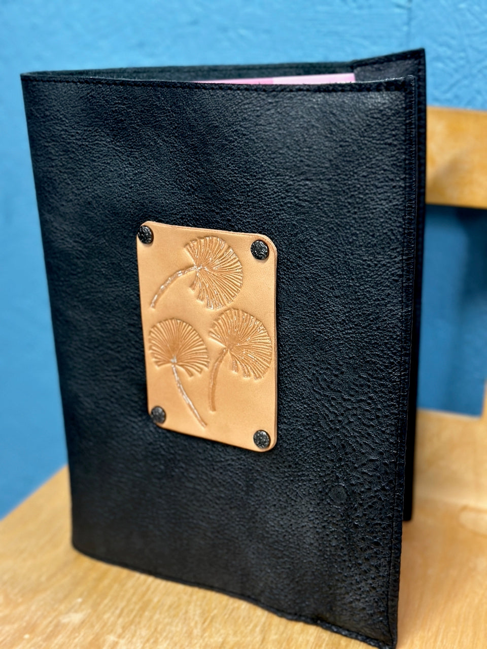 Black leather notebook with a tan leather patch featuring floral gingko leaf embossing on a blue background.