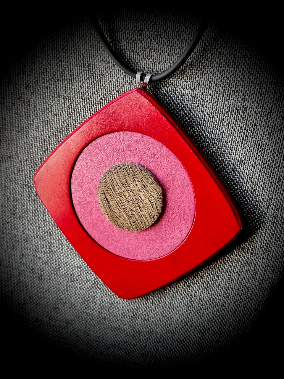 Necklace with a red square pendant featuring a pink circle and hair on hide brown leather center on a gray textured background