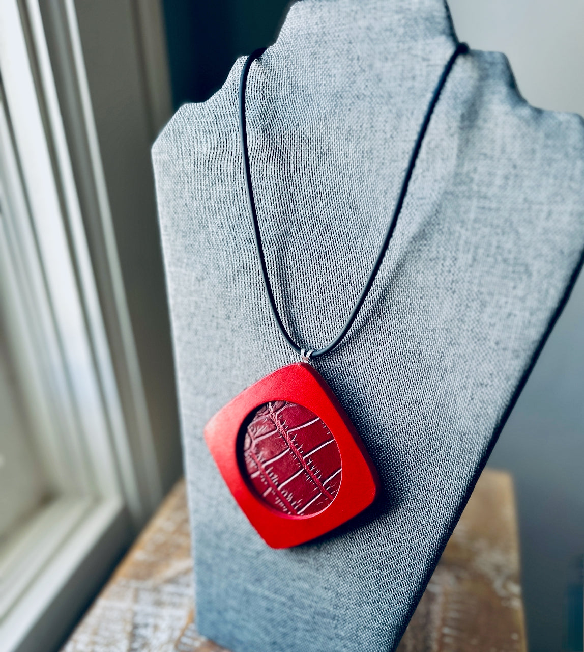 Red leather pendant necklace on a gray fabric easel, made by Undercover Popular Leather Goods